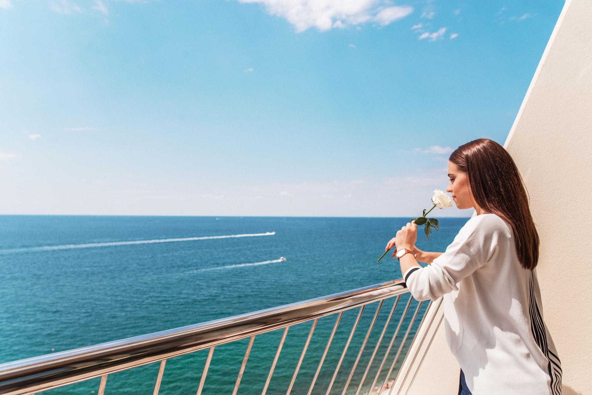 Grand Hotel Bernardin Portoroz Exterior photo The photo depicts a woman standing on a balcony overlooking the ocean. She holds a white rose and looks thoughtfully toward the water, which appears calm and blue. There are two boats visible on the surface of the sea, creating gentle waves. The sky 