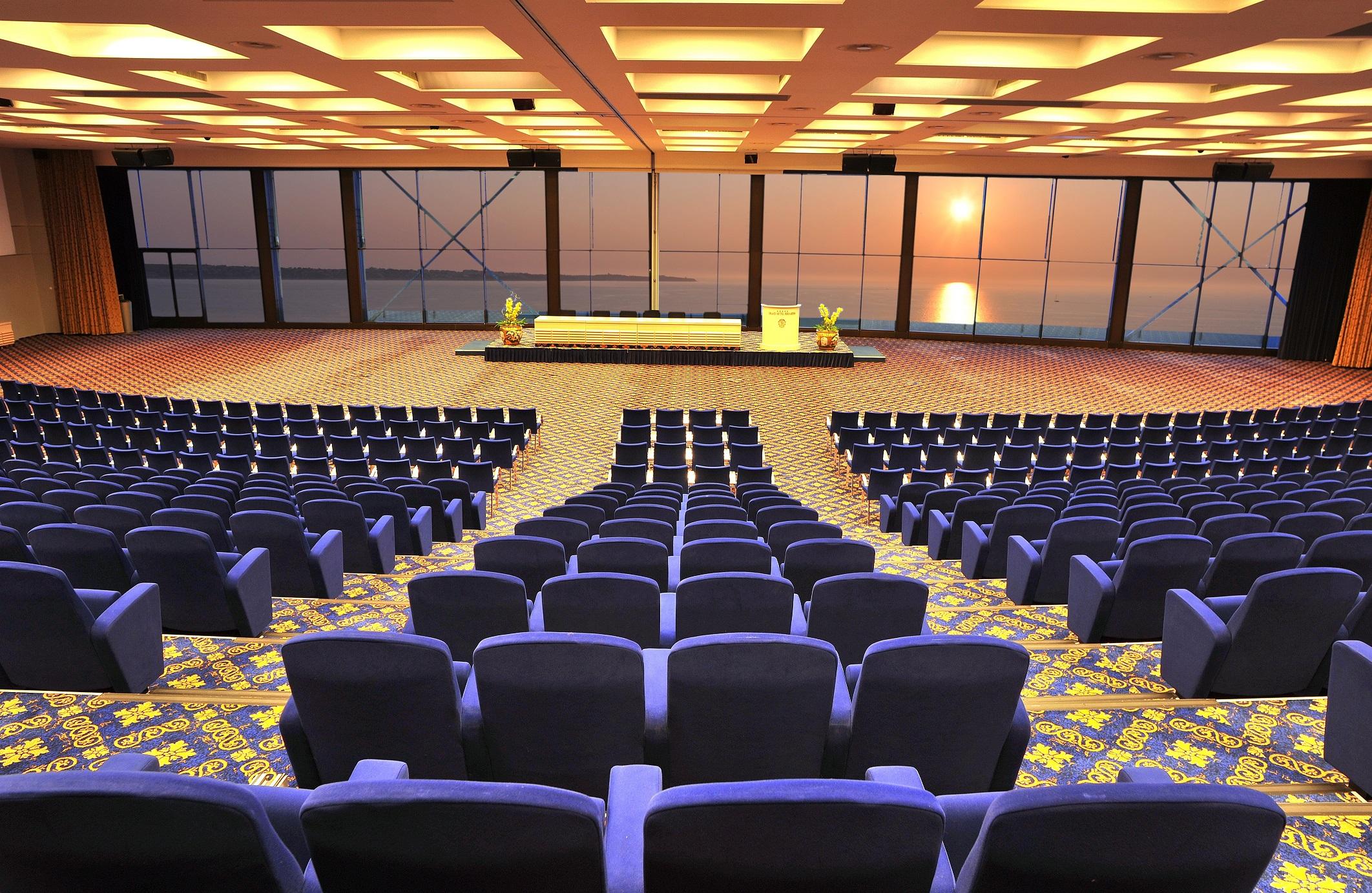 Grand Hotel Bernardin Portoroz Exterior photo The photo shows a spacious conference room or auditorium with a large seating area. The seats are arranged in rows, and they are upholstered in a blue fabric. In the background, there are large windows that provide a view of the sea and a sunset or s