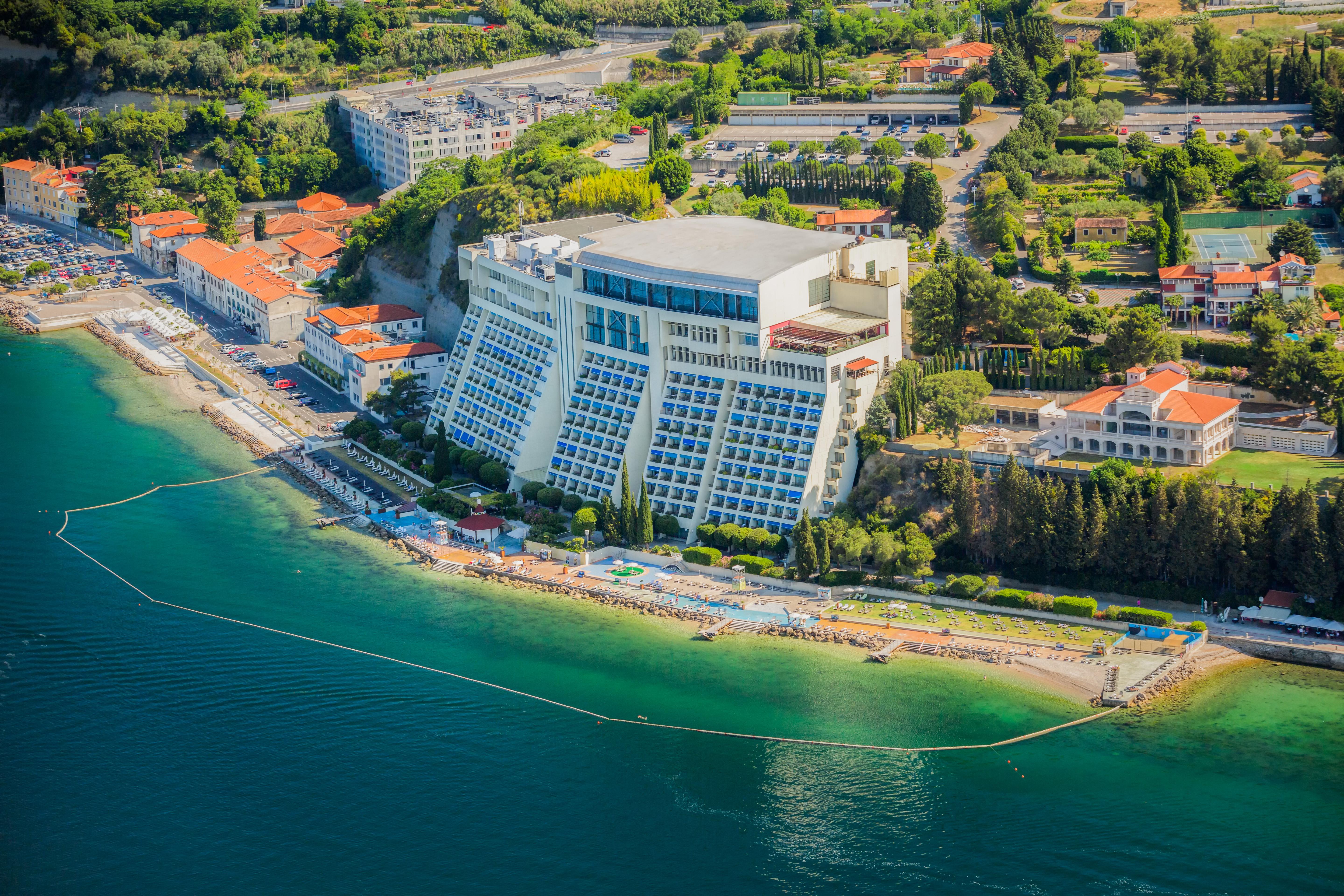 Grand Hotel Bernardin Portoroz Exterior photo The photo shows an aerial view of a coastal area featuring a large hotel building situated on a beach. The hotel has multiple floors and appears to have a modern design, with a structured façade facing the water. Surrounding the hotel are various bui