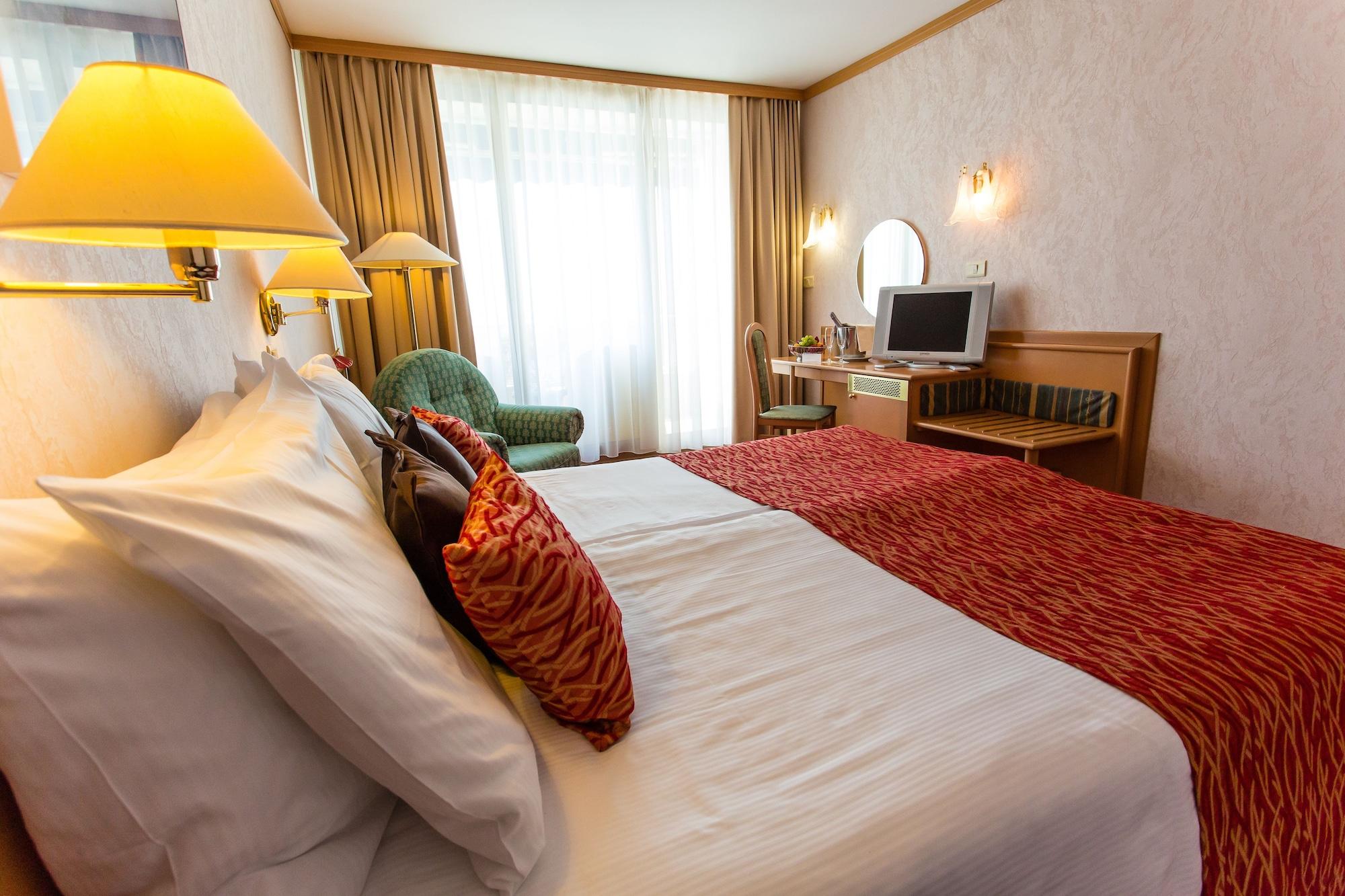 Grand Hotel Bernardin Portoroz Exterior photo The photo shows a hotel room featuring two beds, each with decorative pillows. There is a warm color scheme, highlighted by a red bedspread. To the side, a green armchair is visible, along with a desk that has a mirror and a television on it. Large w