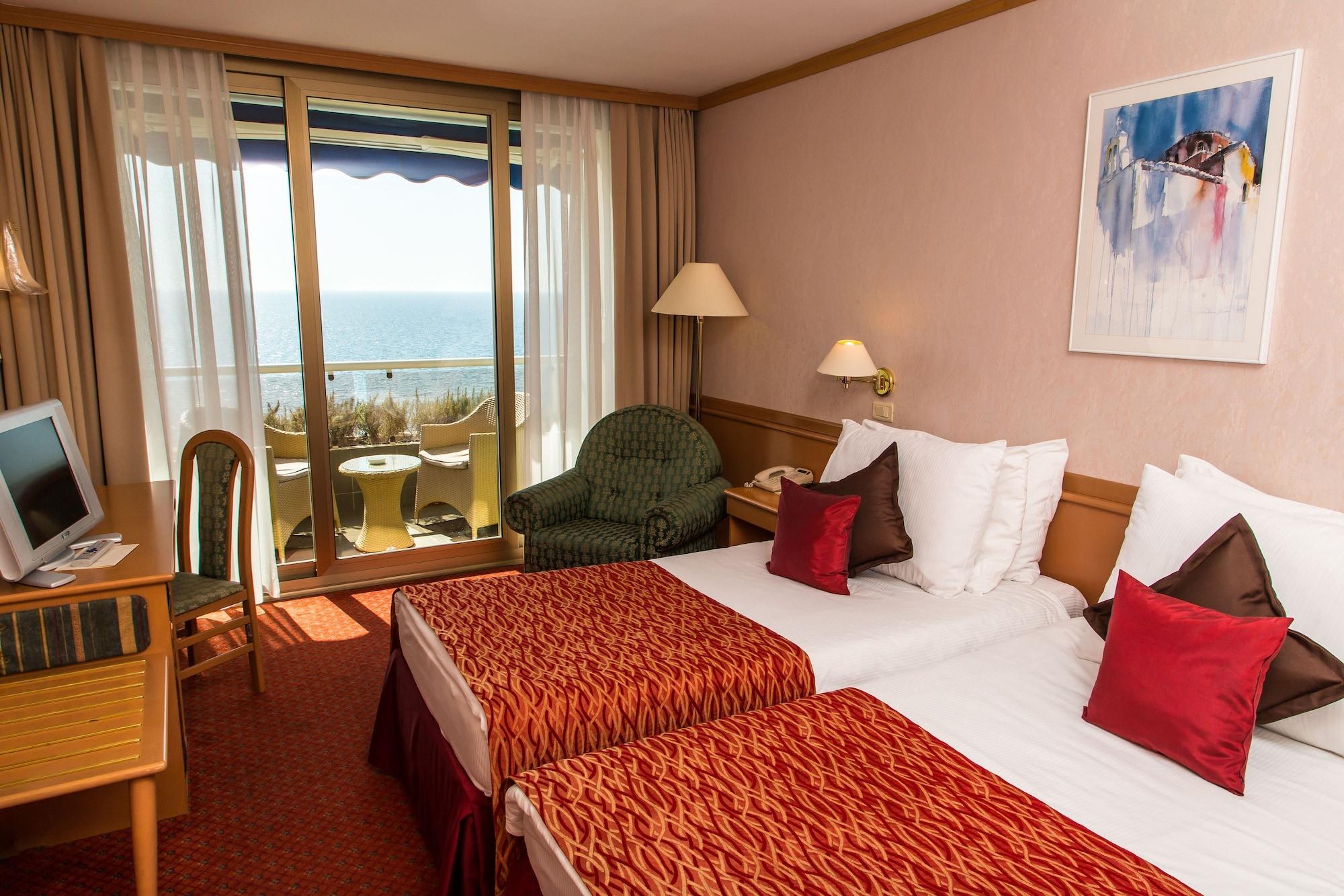 Grand Hotel Bernardin Portoroz Exterior photo The photo shows a hotel room featuring two twin beds with decorative red and white bedding. There is a small seating area with a green chair and a side table. The room has large windows that offer a view of the ocean, and a balcony with two chairs is