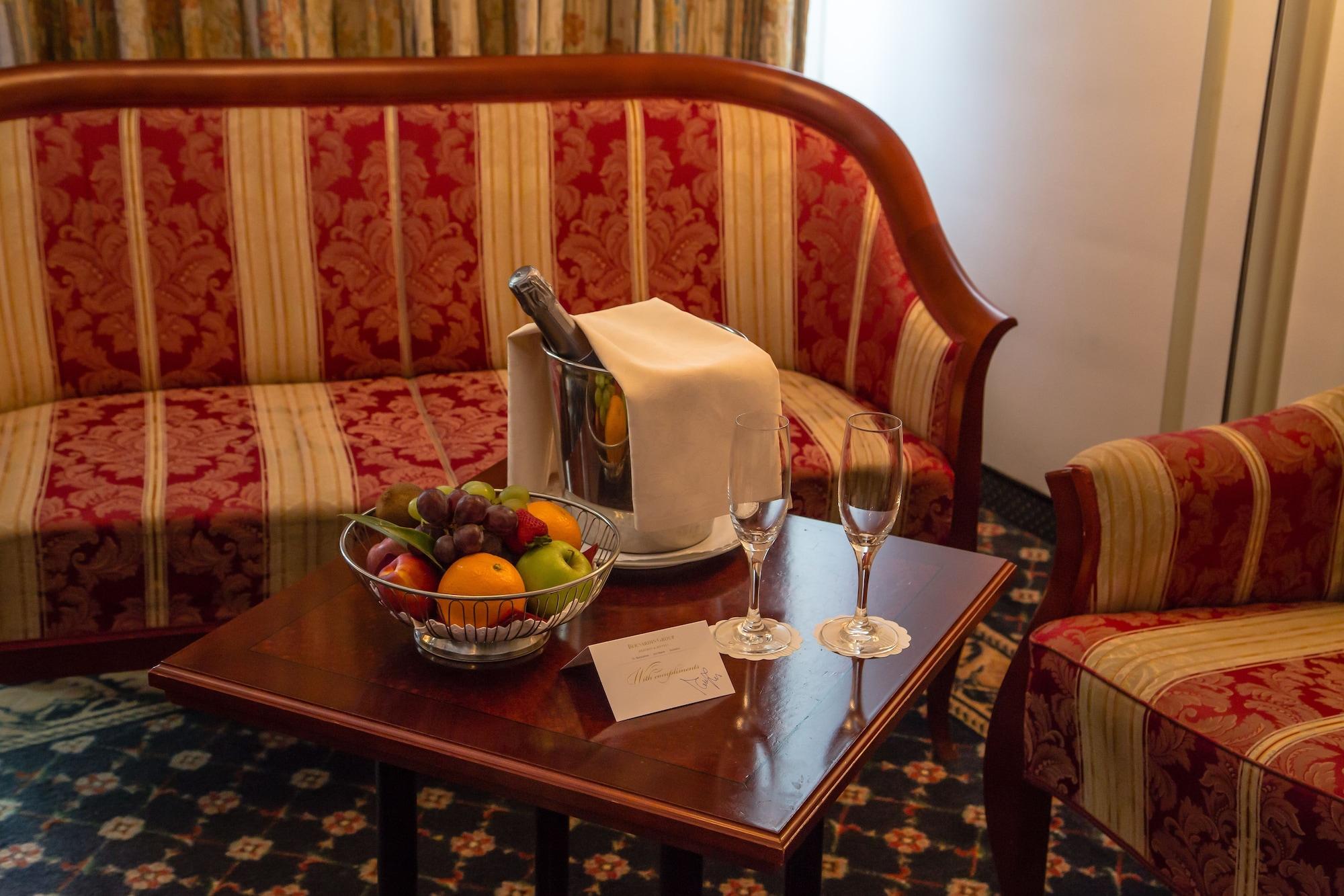 Grand Hotel Bernardin Portoroz Exterior photo The photo depicts a cozy hotel room setting. In the center, there is a small wooden table adorned with a silver champagne bucket, a white cloth draped over it, and two champagne flutes. Next to it, there is a bowl of assorted fruits, including orange