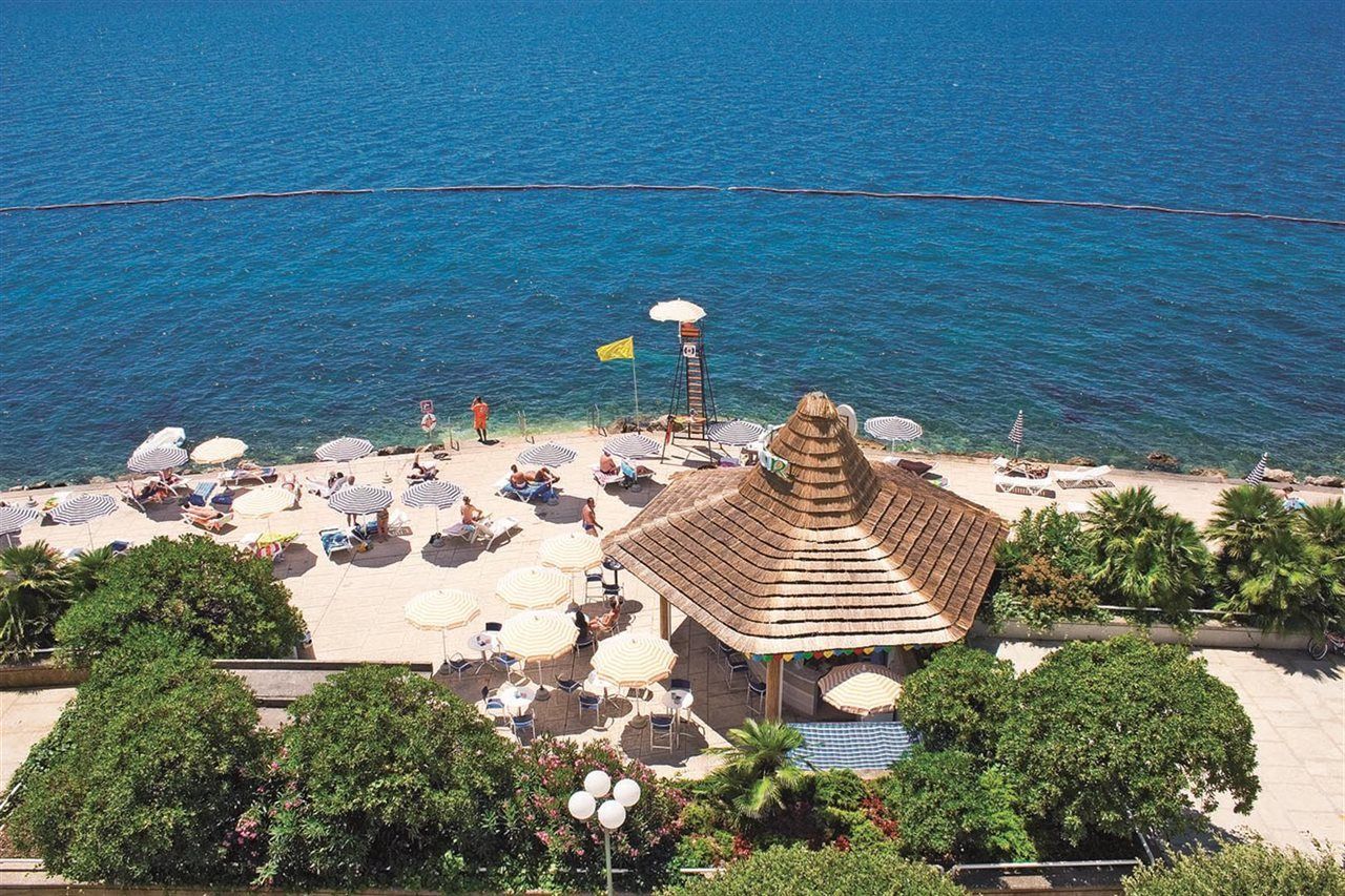 Grand Hotel Bernardin Portoroz Exterior photo The photo shows a picturesque beach scene with clear blue water. There is a sandy area with several white umbrellas and lounge chairs where people are relaxing. A small thatched-roof gazebo or hut is visible, likely serving as a food or drink stand. 