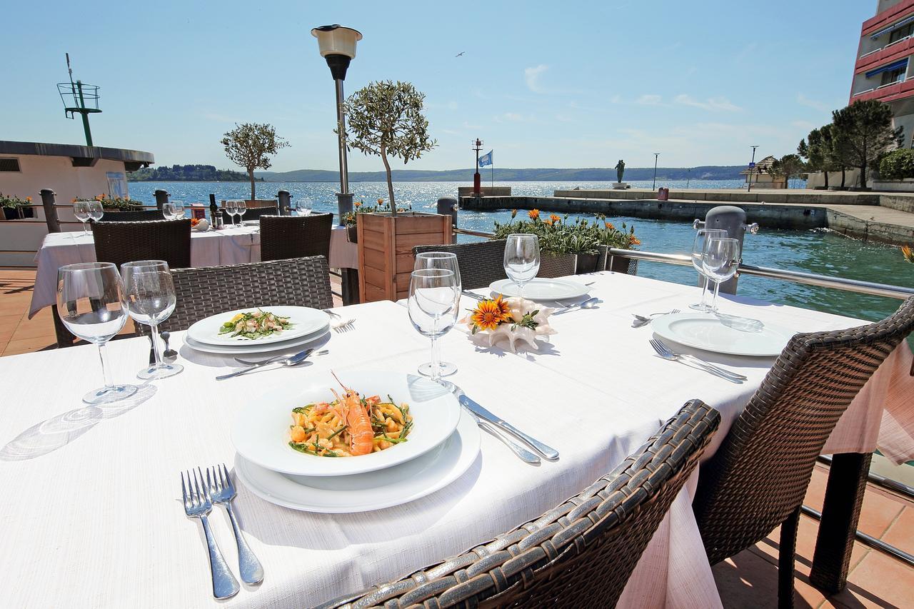 Grand Hotel Bernardin Portoroz Exterior photo The photo shows a picturesque outdoor dining setup by a waterfront. There are several tables with elegant white tablecloths, each set with plates, cutlery, and wine glasses. In the foreground, a plate of pasta with salmon is prominently displayed alo