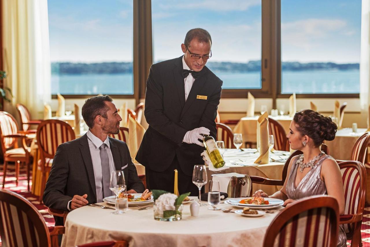 Grand Hotel Bernardin Portoroz Exterior photo The image shows a fine dining setting in a restaurant. In the foreground, a well-dressed couple is seated at an elegantly set table. The man is looking towards the waiter, who is pouring a drink from a bottle into a wine glass. The woman is sitting b