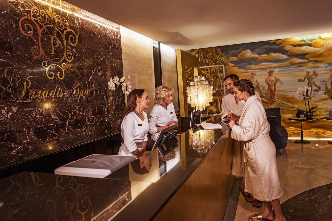 Grand Hotel Bernardin Portoroz Exterior photo The photo shows a luxurious spa reception area. There are three staff members behind a sleek, dark marble reception desk, wearing white robes. Two guests, also in robes, are interacting with the staff. The background features an elegant mural and sty