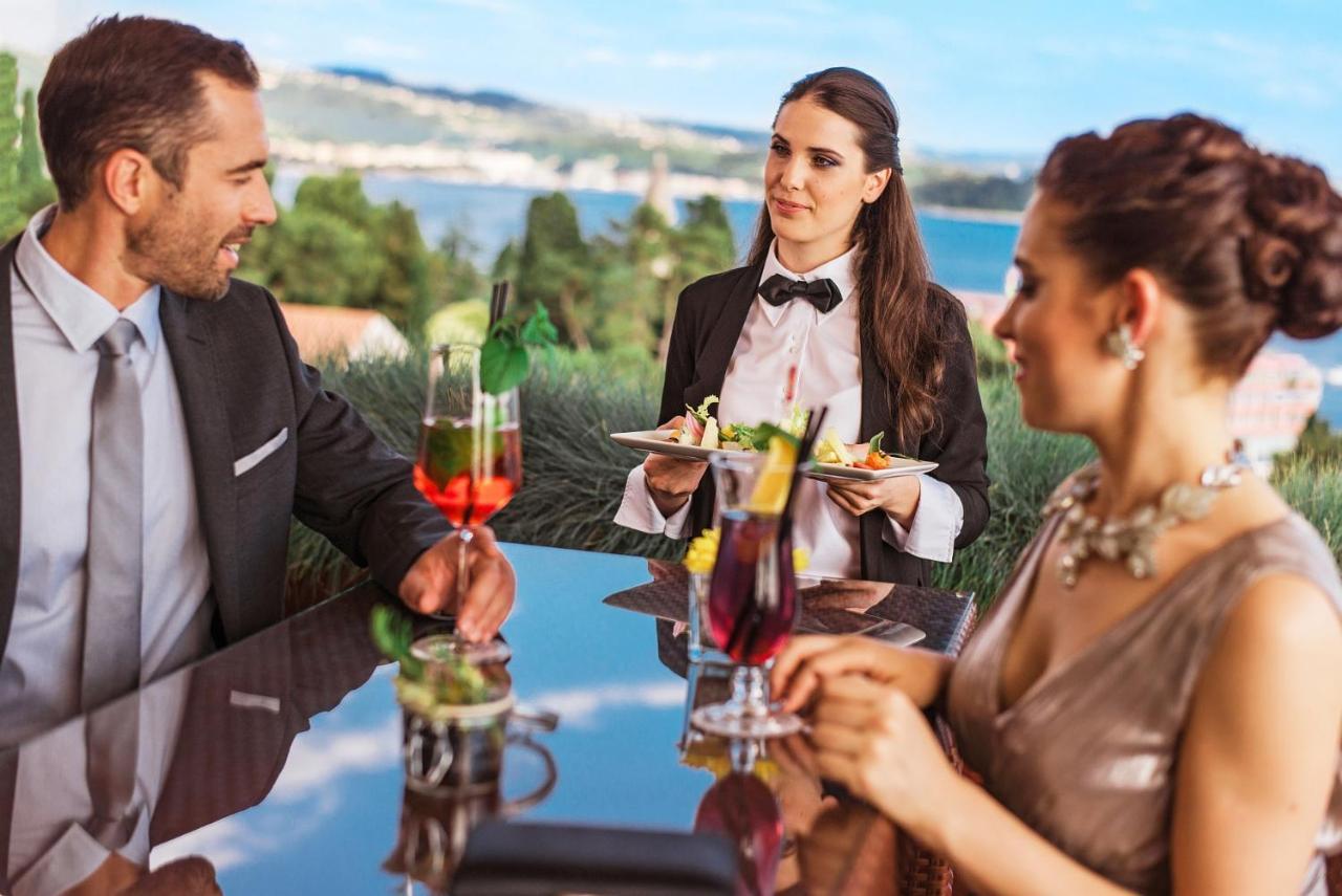 Grand Hotel Bernardin Portoroz Exterior photo The photo shows a dining scene likely set in a restaurant with a picturesque view. In the image, a man and a woman are seated at a table, both dressed elegantly. The woman is wearing a sparkling dress, and the man is in a formal suit. A waitress, dre