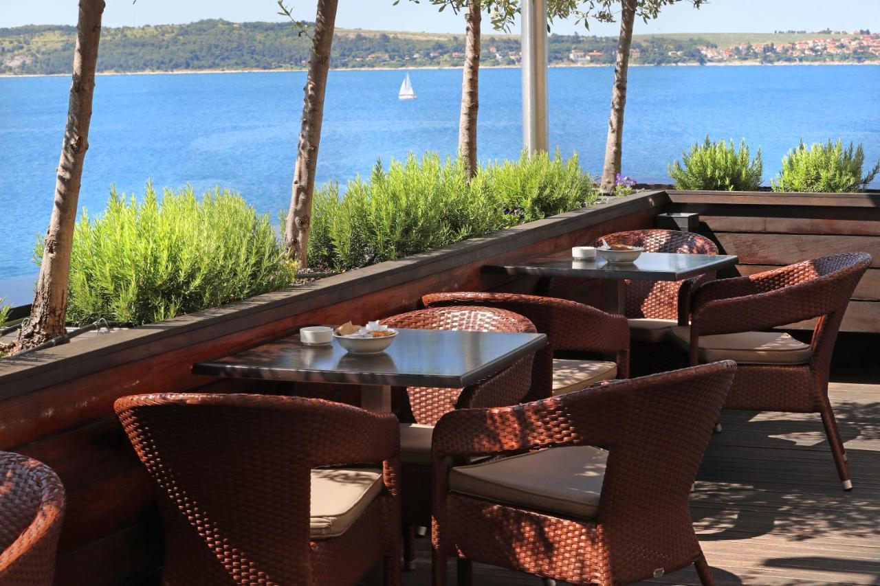 Grand Hotel Bernardin Portoroz Exterior photo The photo shows an outdoor seating area of a restaurant or café with several wicker chairs and tables. The setting is surrounded by green plants and has a view of a calm body of water, likely a lake or sea, in the background. In the distance, there's