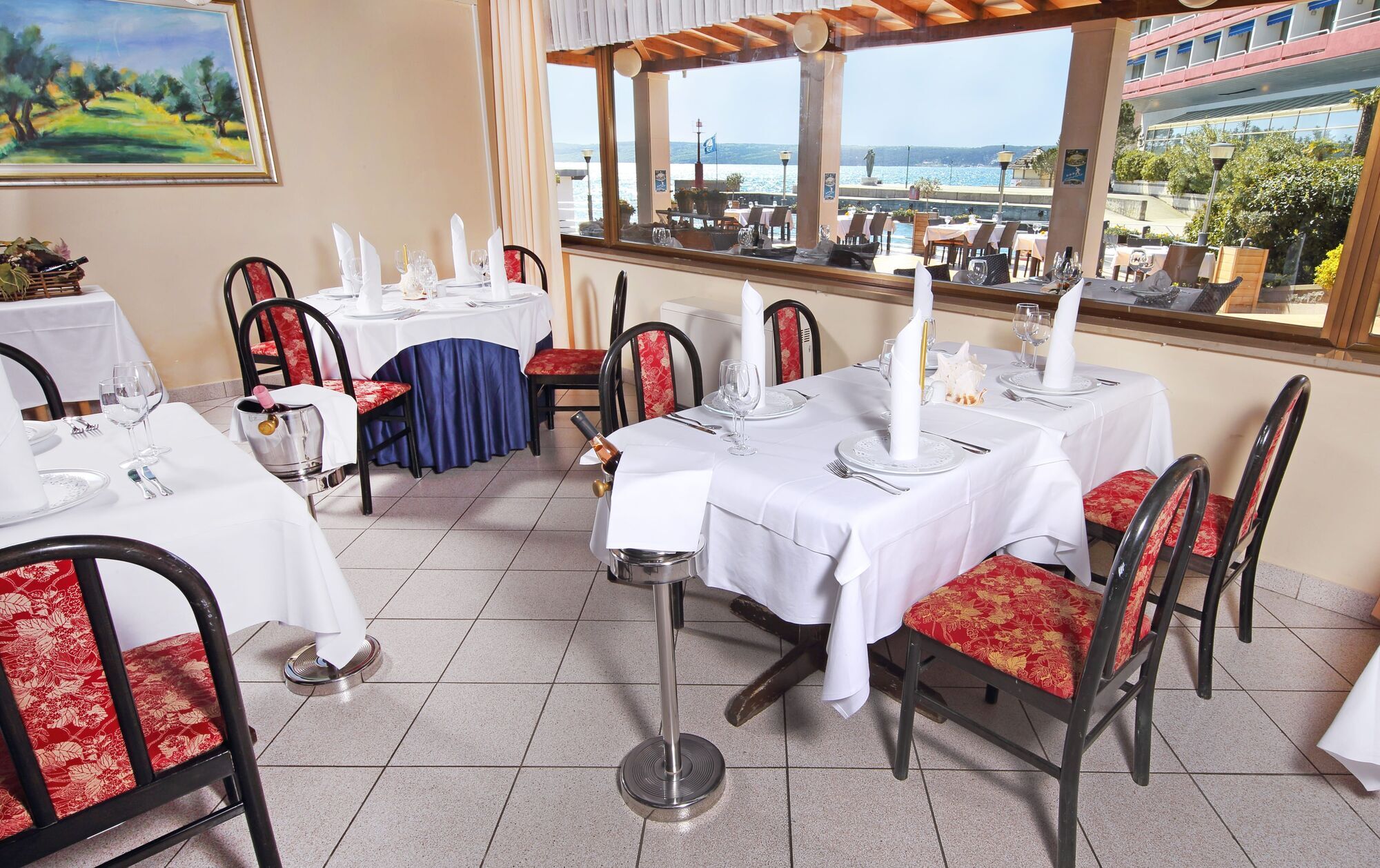 Grand Hotel Bernardin Portoroz Exterior photo The photo shows the interior of a dining area in a restaurant. There are several tables, each covered with white tablecloths. The chairs are a mix of black frames with either floral or solid-colored cushions. Large windows provide a view of the outsi