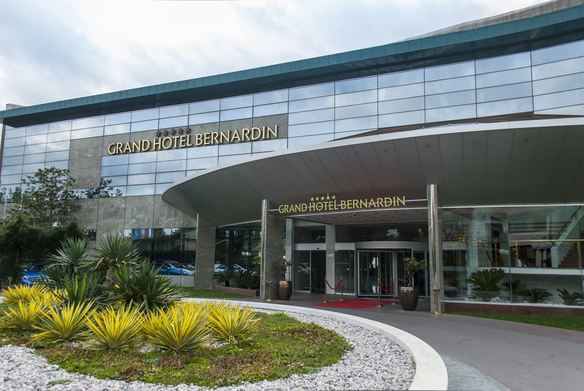 Grand Hotel Bernardin Portoroz Exterior photo The photo showcases the entrance of the Grand Hotel Bernardin, featuring a modern architectural design. The facade includes large glass windows and a sleek overhang. Strategically placed greenery, such as palms and low shrubs, adds to the aesthetic a